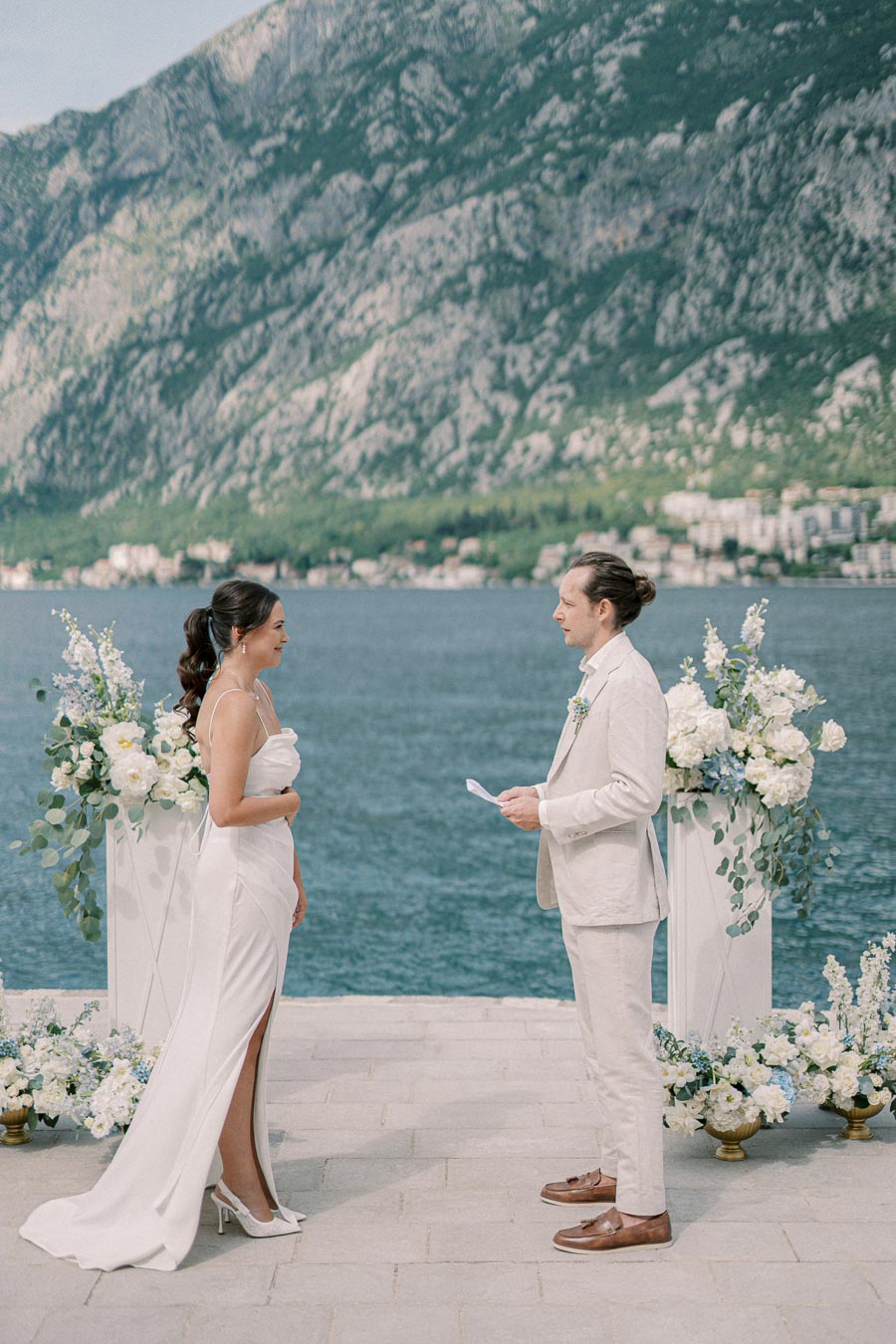A couple exchanges vows in an elegant outdoor wedding ceremony by a picturesque lake, with a stunning mountainous backdrop. Lush floral arrangements in white and pastel colors adorn the scene.