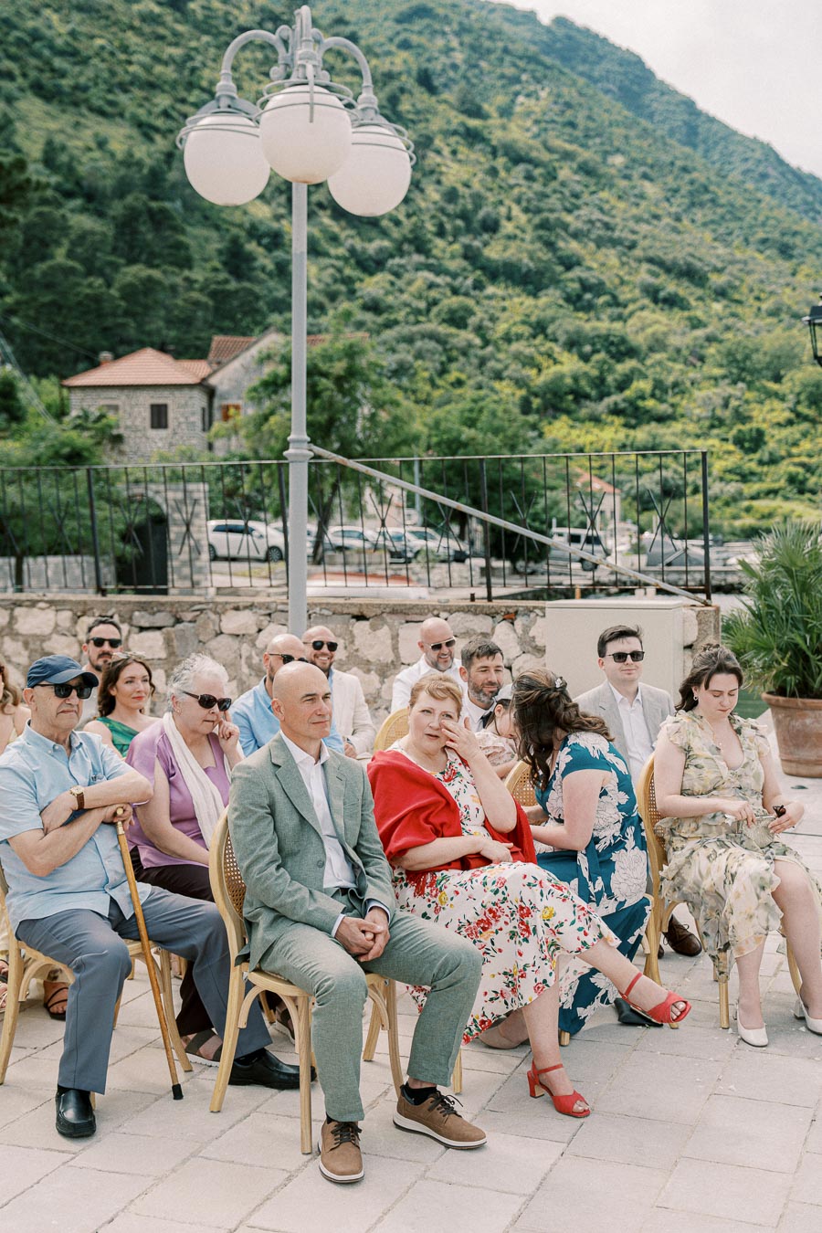 A group of people sitting on outdoor chairs in a scenic, mountainous location with a stone building and lush greenery in the background. They appear to be dressed for a formal occasion. A vintage-style lamp post is visible nearby.