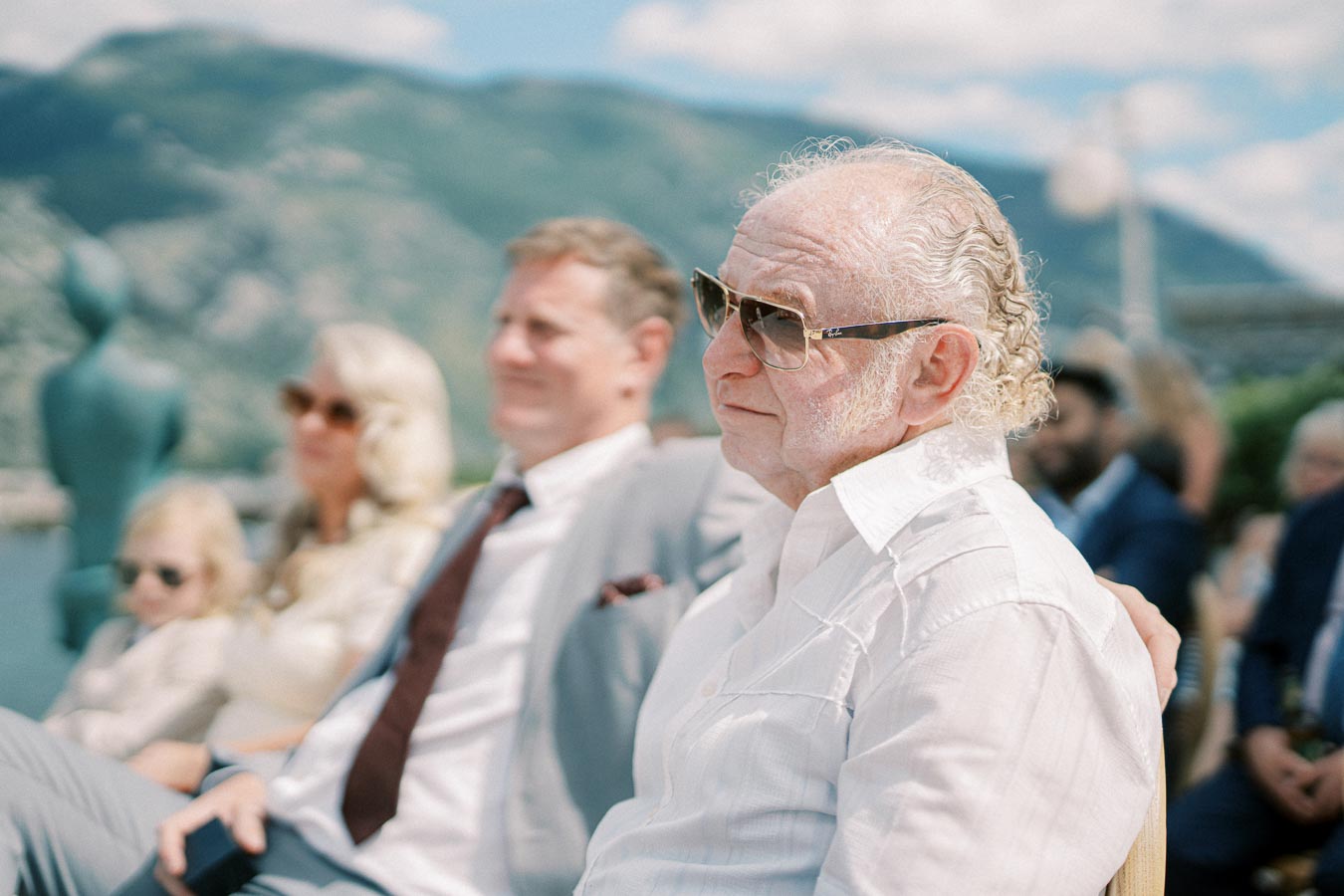 Elderly man in sunglasses and white shirt sitting outdoors at an event, with blurred people and mountainous landscape in the background.