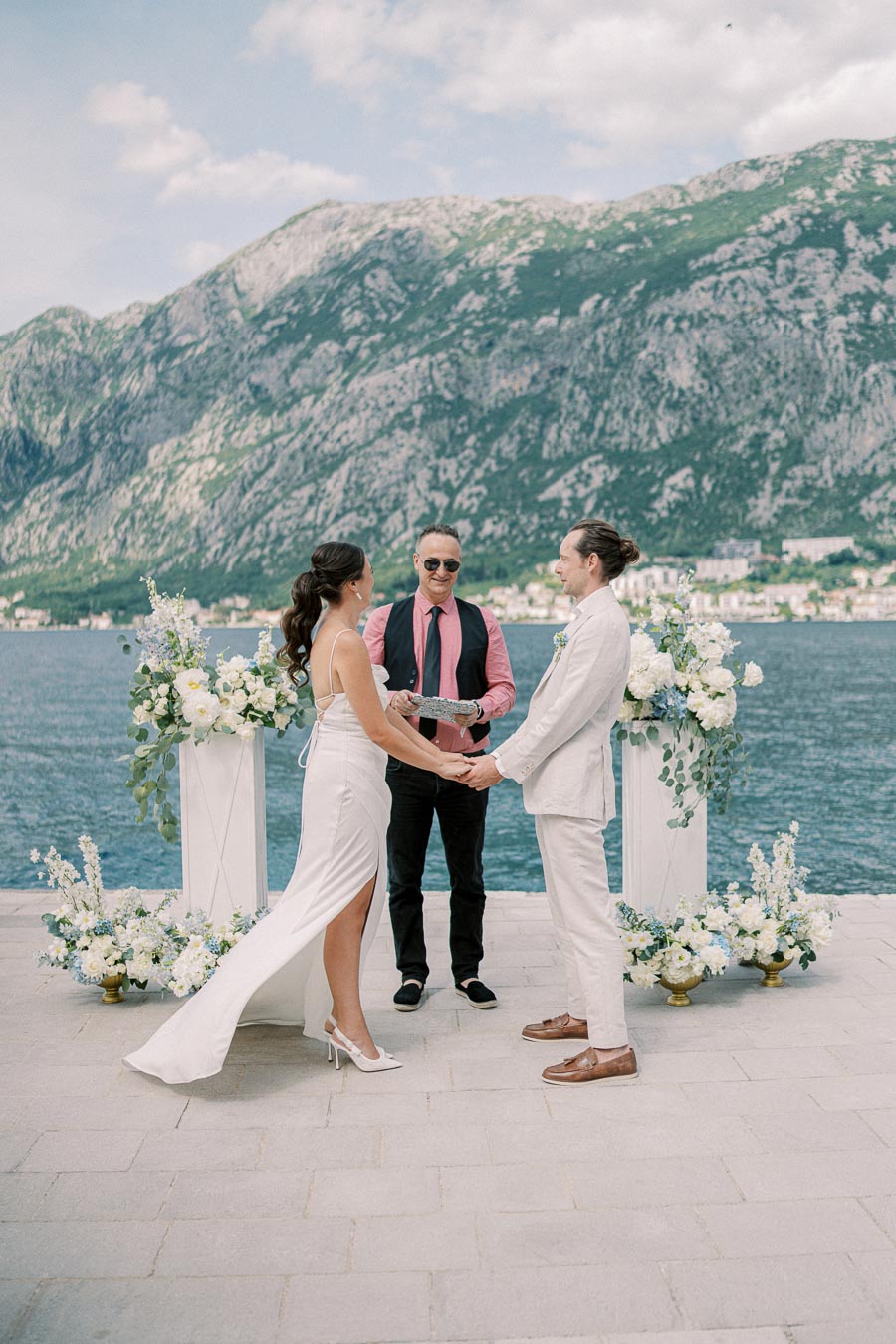 Elegant outdoor wedding ceremony by a scenic lakeside with majestic mountain backdrop, featuring a couple holding hands, exchanging vows with a celebrant, surrounded by beautiful floral arrangements.