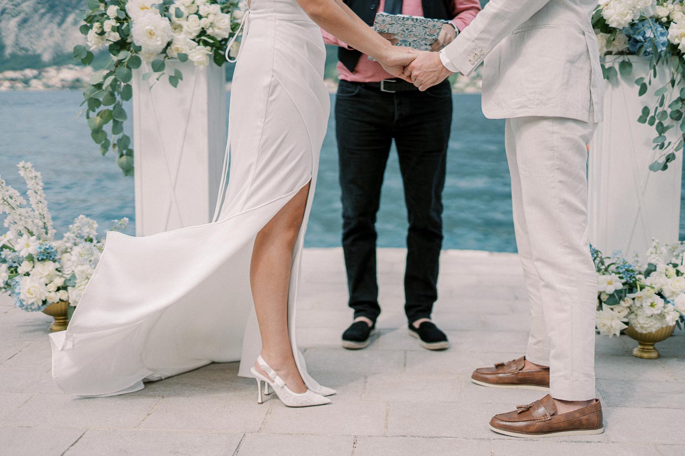 Elegant wedding ceremony by the waterfront, featuring a couple in stylish attire holding hands with a scenic ocean backdrop and floral arrangements.