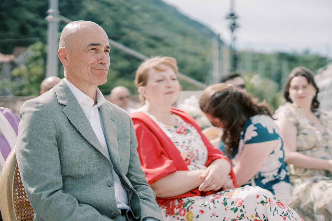 A group of people sitting outdoors, dressed in formal attire, with a scenic mountain backdrop. In the foreground, a man in a gray suit and a woman in a floral dress with a red cardigan are seated and appear engaged in an outdoor event under sunny skies.