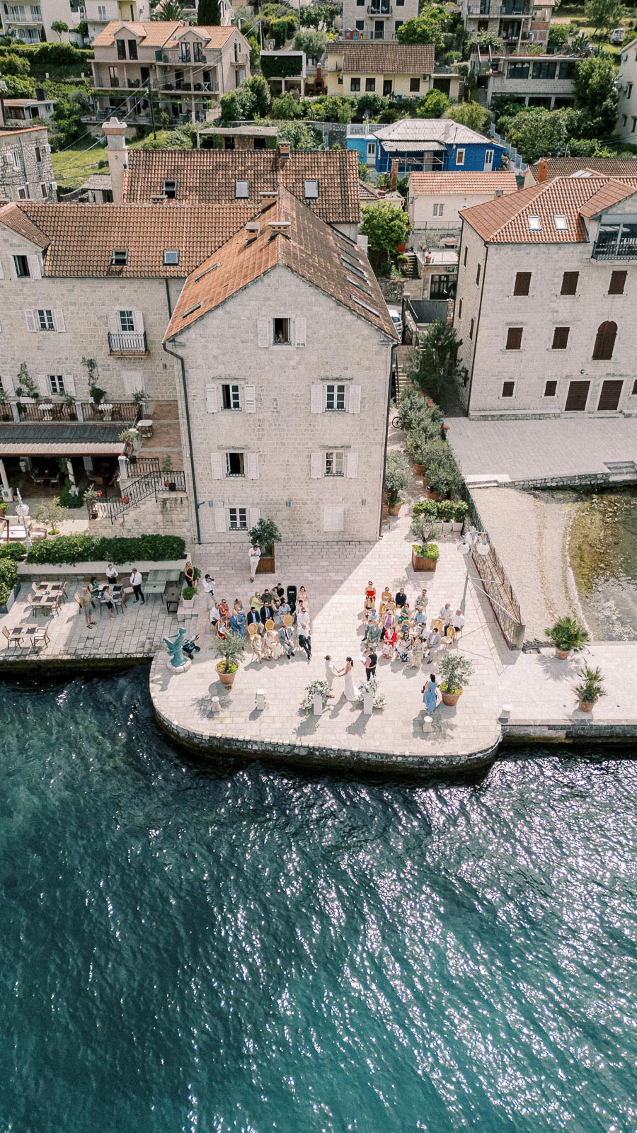 Aerial view of an outdoor wedding ceremony by the water, surrounded by historic stone buildings with terracotta roofs, featuring guests seated on a stone courtyard overlooking the sea.