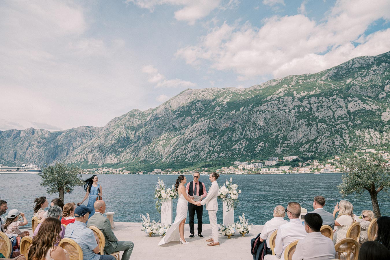 Outdoor wedding ceremony by the sea, with a couple holding hands in front of a celebrant. Guests are seated on the patio overlooking a scenic backdrop of mountains and a calm waterfront, surrounded by floral decorations.