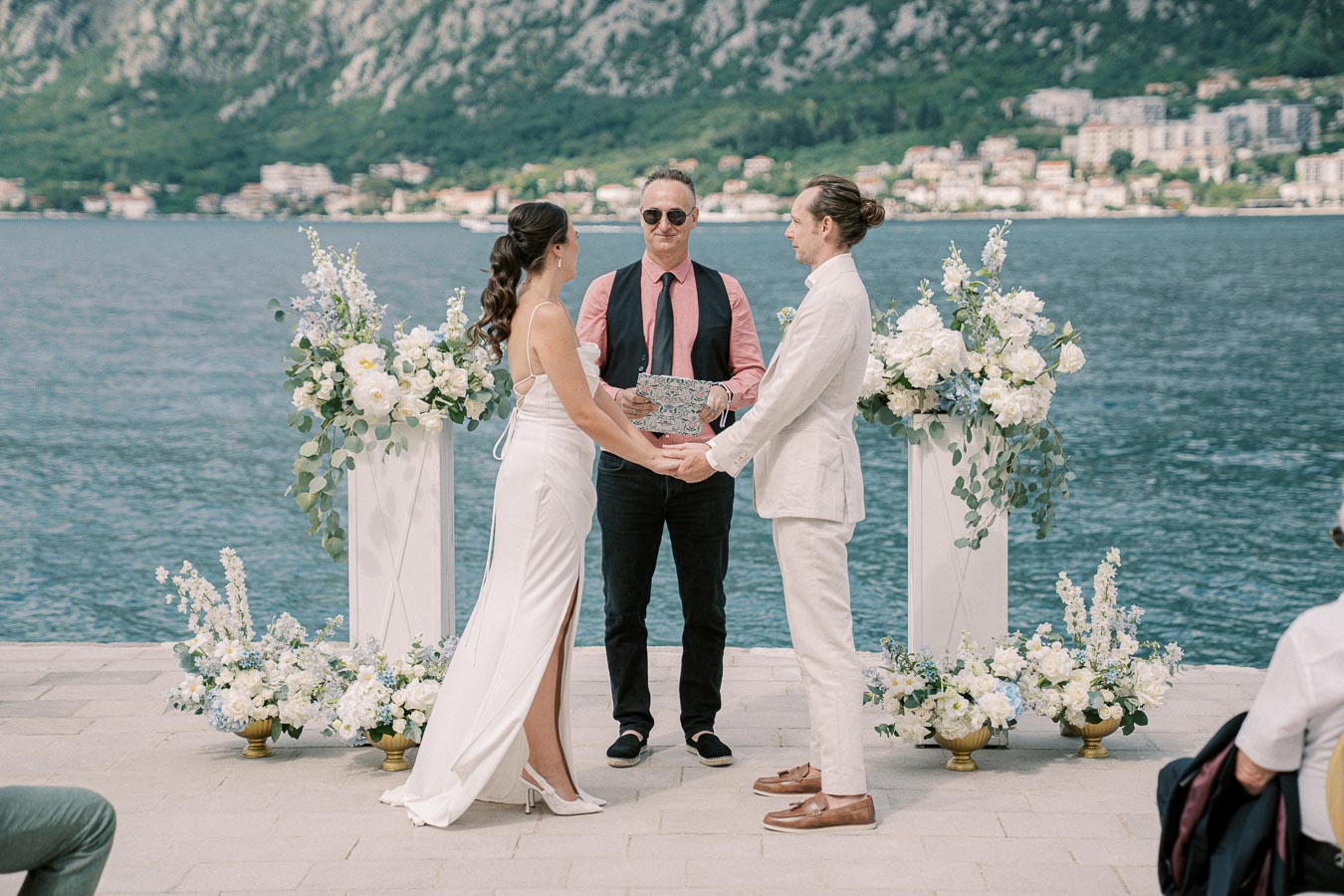 Beachfront wedding ceremony scene with couple holding hands and officiant, surrounded by white floral arrangements, against a backdrop of scenic mountains and water.