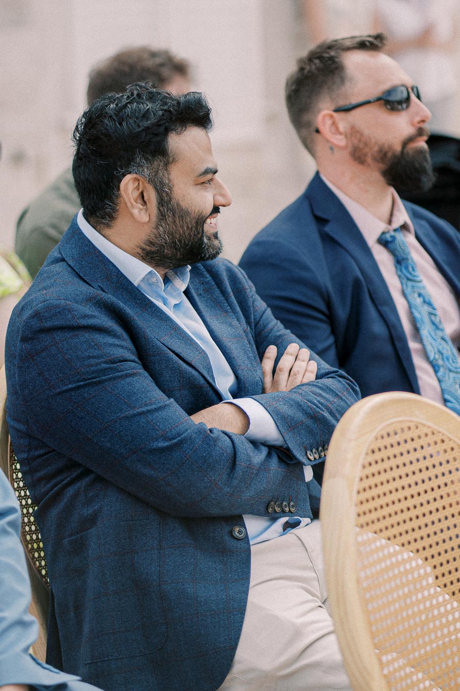 A man in a blue blazer smiling and sitting with arms crossed, attending an outdoor event alongside another man in sunglasses.