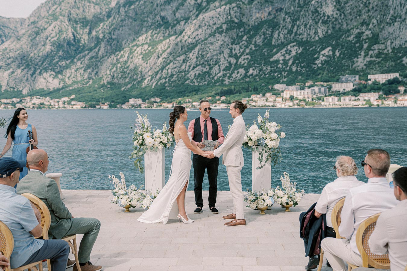 A couple exchanging vows during a picturesque outdoor wedding ceremony by the sea, with a scenic backdrop of mountains and greenery, surrounded by elegantly arranged white floral decorations and seated guests.