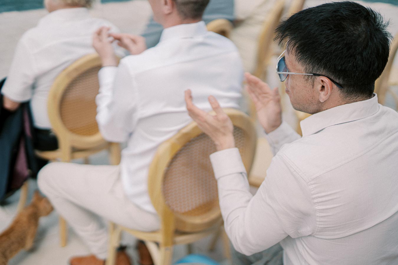 A group of people seated at an outdoor event, with one person clapping and wearing sunglasses, captured from behind. A small cat is visible on the ground near the chairs.