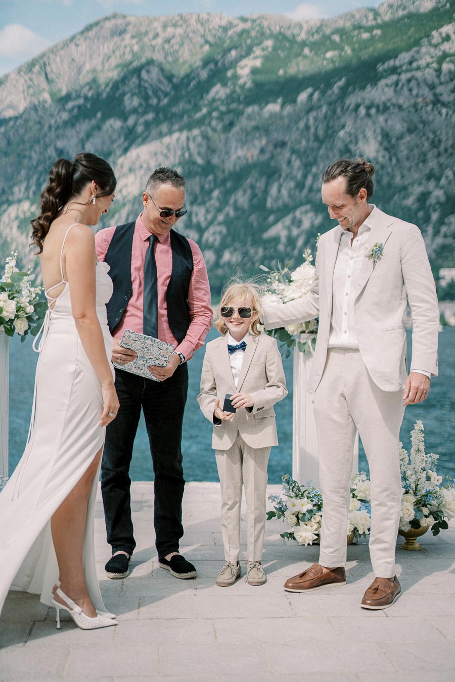 Outdoor wedding ceremony by a scenic lake with mountains in the background, featuring a bride in a white gown, groom in a light suit, young boy in a suit and sunglasses, and an officiant with sunglasses holding a book.