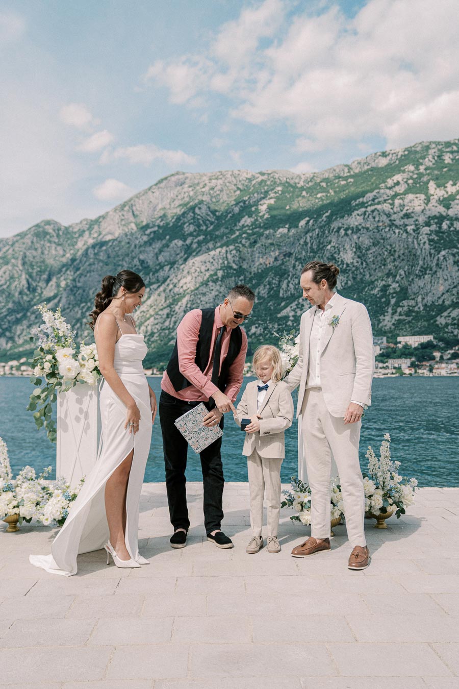 A wedding ceremony by the sea with a bride in a white gown and groom in a beige suit, accompanied by a child in a suit and a man officiating. Scenic mountainous backdrop under a bright, cloudy sky.