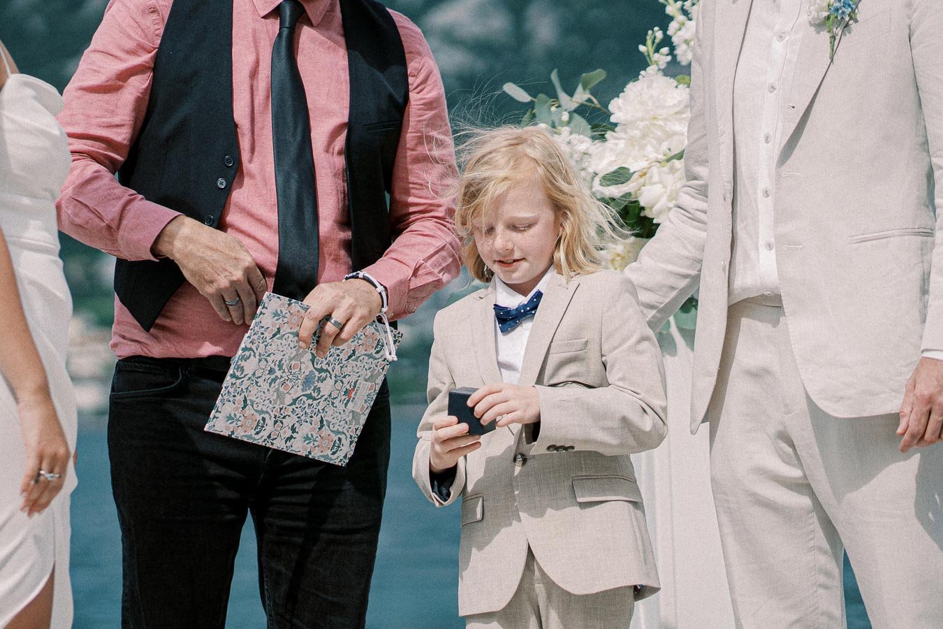 Young boy in a beige suit holds a small object during an outdoor wedding ceremony, standing between two adults, one in black vest and pink shirt, another in a beige suit.