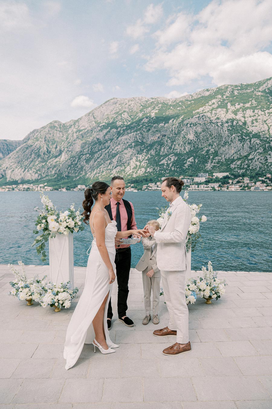 A couple exchanging rings during a waterfront wedding ceremony with scenic mountain views in the background.
