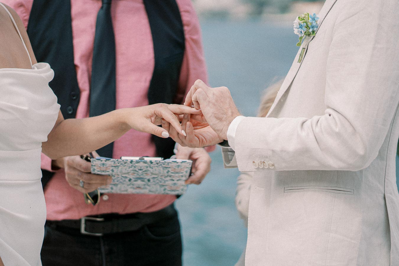 A bride and groom exchange rings during a waterfront wedding ceremony, with an officiant holding ceremony notes in the background.