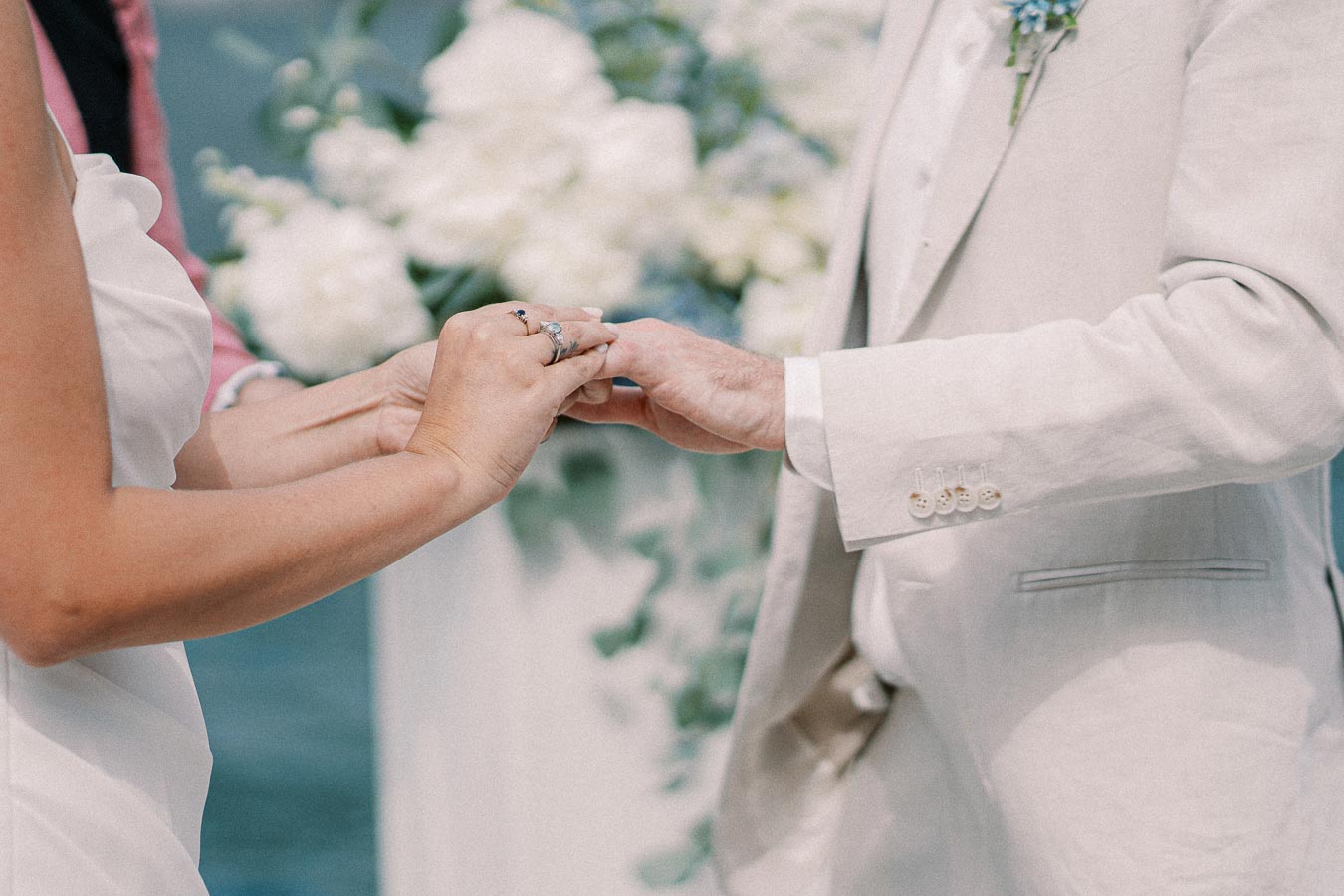 Close-up of a wedding couple exchanging rings during their outdoor ceremony, with the groom wearing a light suit and the bride in a white dress, surrounded by floral decorations.