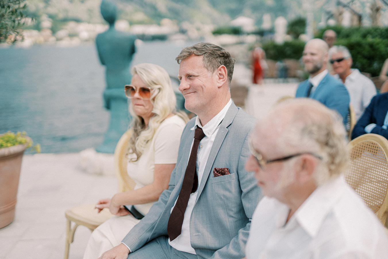 A group of people sitting outdoors at an elegant gathering near a waterfront, with one man in focus wearing a light blue suit and smiling. The background features a scenic view of the water and distant greenery under a clear sky.