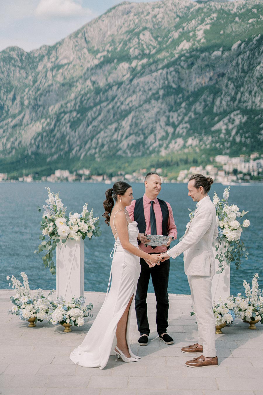 A couple exchanging vows in an outdoor wedding ceremony by a picturesque lake, surrounded by lush mountains and floral arrangements, under a clear blue sky.