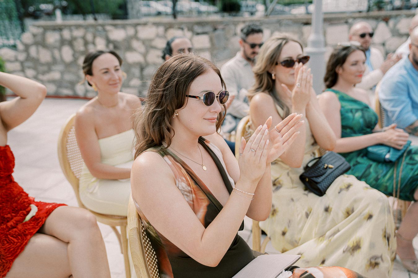 Audience at an outdoor event seated on wicker chairs, with people applauding and wearing stylish summer dresses and sunglasses, set against a stone wall backdrop.