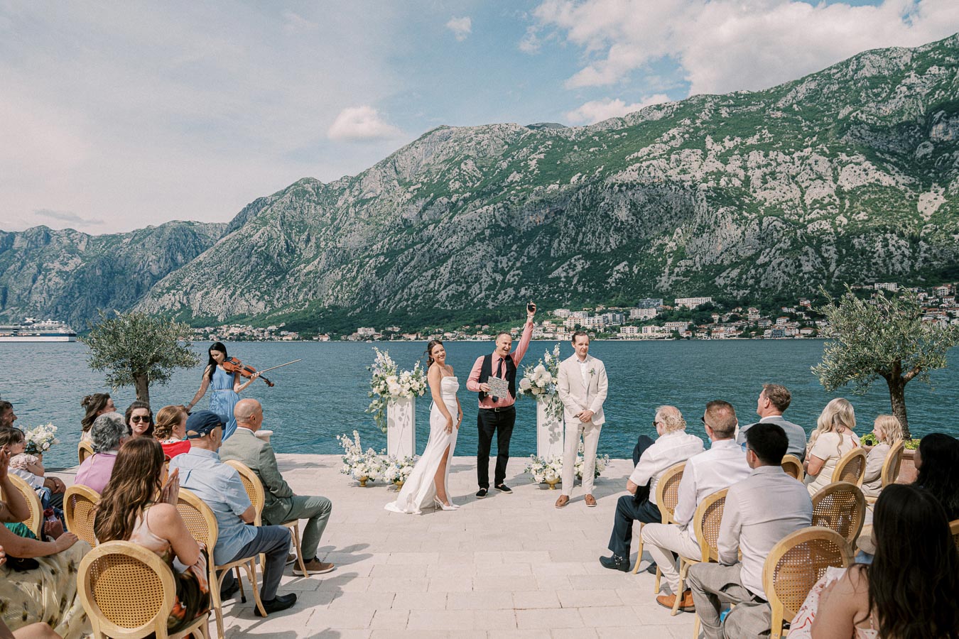 Elegant outdoor wedding ceremony by the sea with mountainous backdrop, featuring seated guests, a couple at the altar, a violinist playing, and officiant joyful.