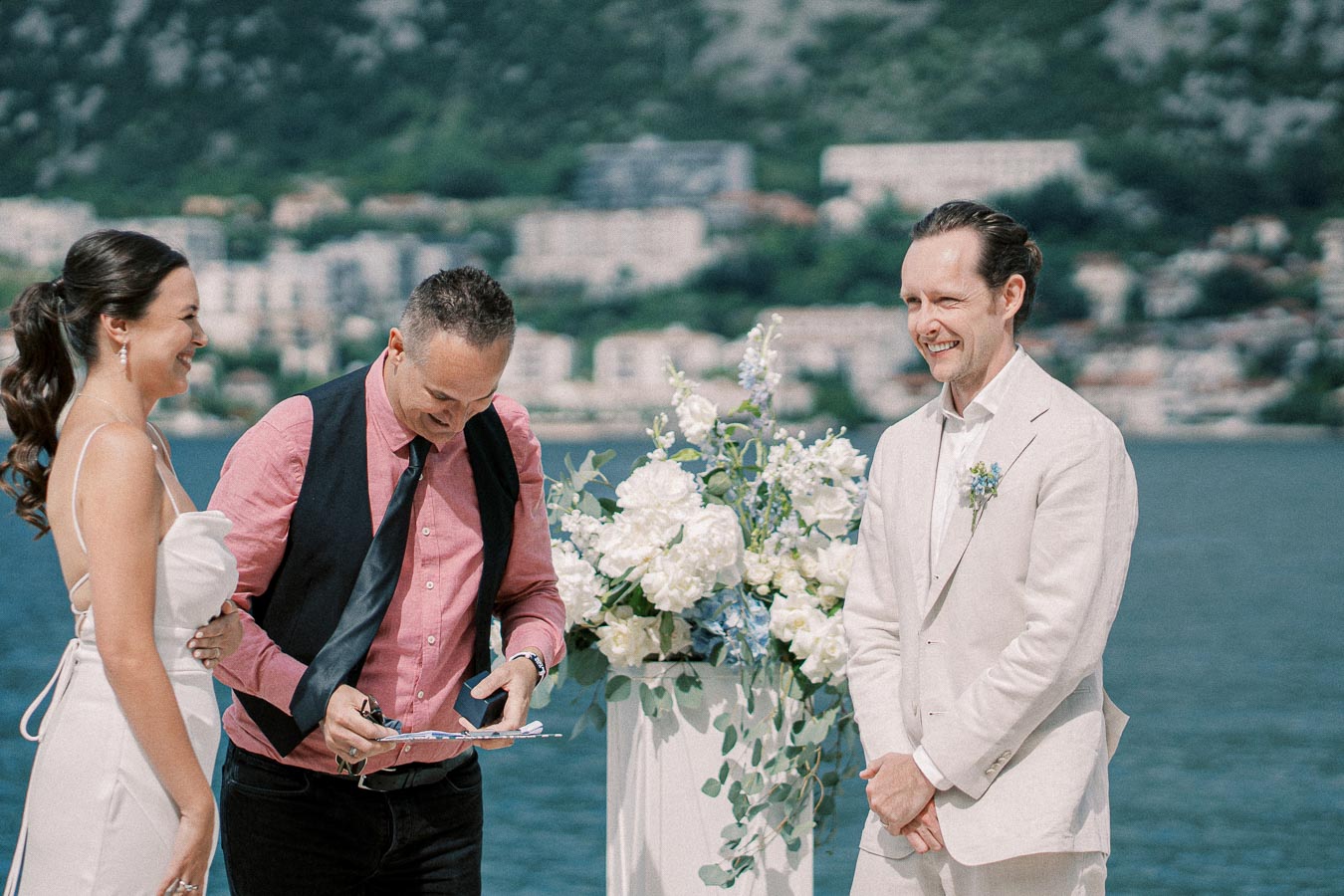 Wedding ceremony by the water with a happy couple and officiant, surrounded by floral arrangements, set against a scenic background of buildings and greenery.