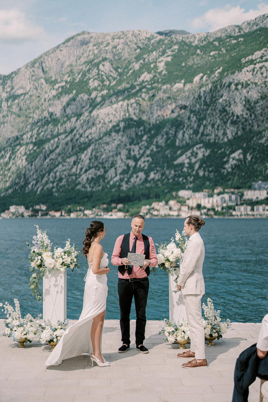 Couple exchanging vows during an outdoor wedding ceremony by the waterfront with mountainous landscape in the background, surrounded by elegant floral arrangements.