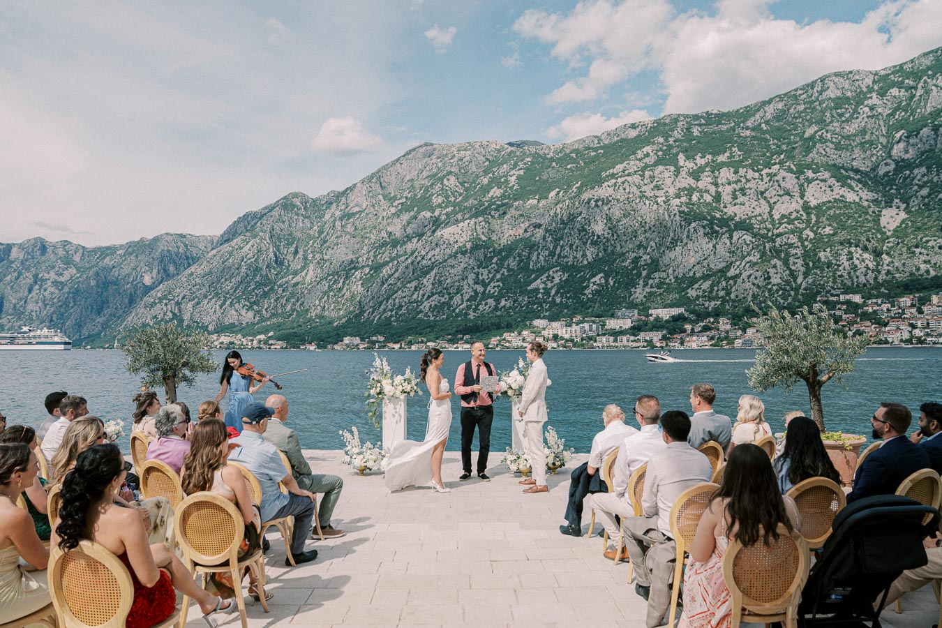 Outdoor wedding ceremony by the scenic bay with mountains in the background, featuring a couple standing at the altar, surrounded by guests seated in chairs.
