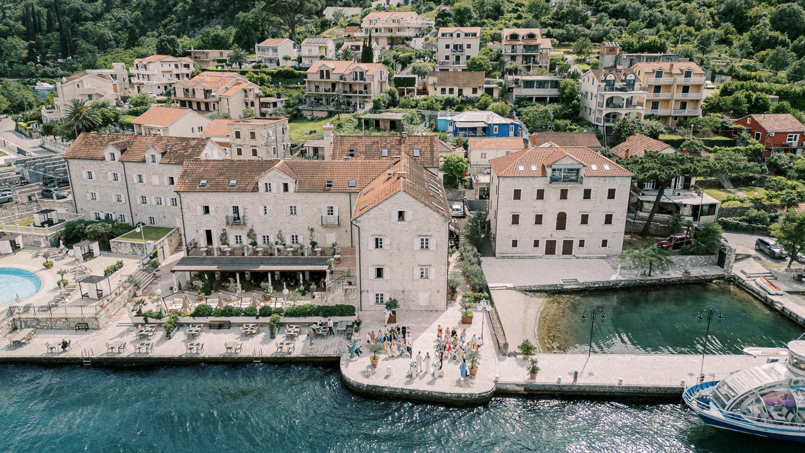 Aerial view of a quaint coastal village with historic stone buildings, surrounded by lush greenery. The image captures a vibrant waterfront scene with a lively gathering of people near a pier, adjacent to the deep blue waters of the ocean.
