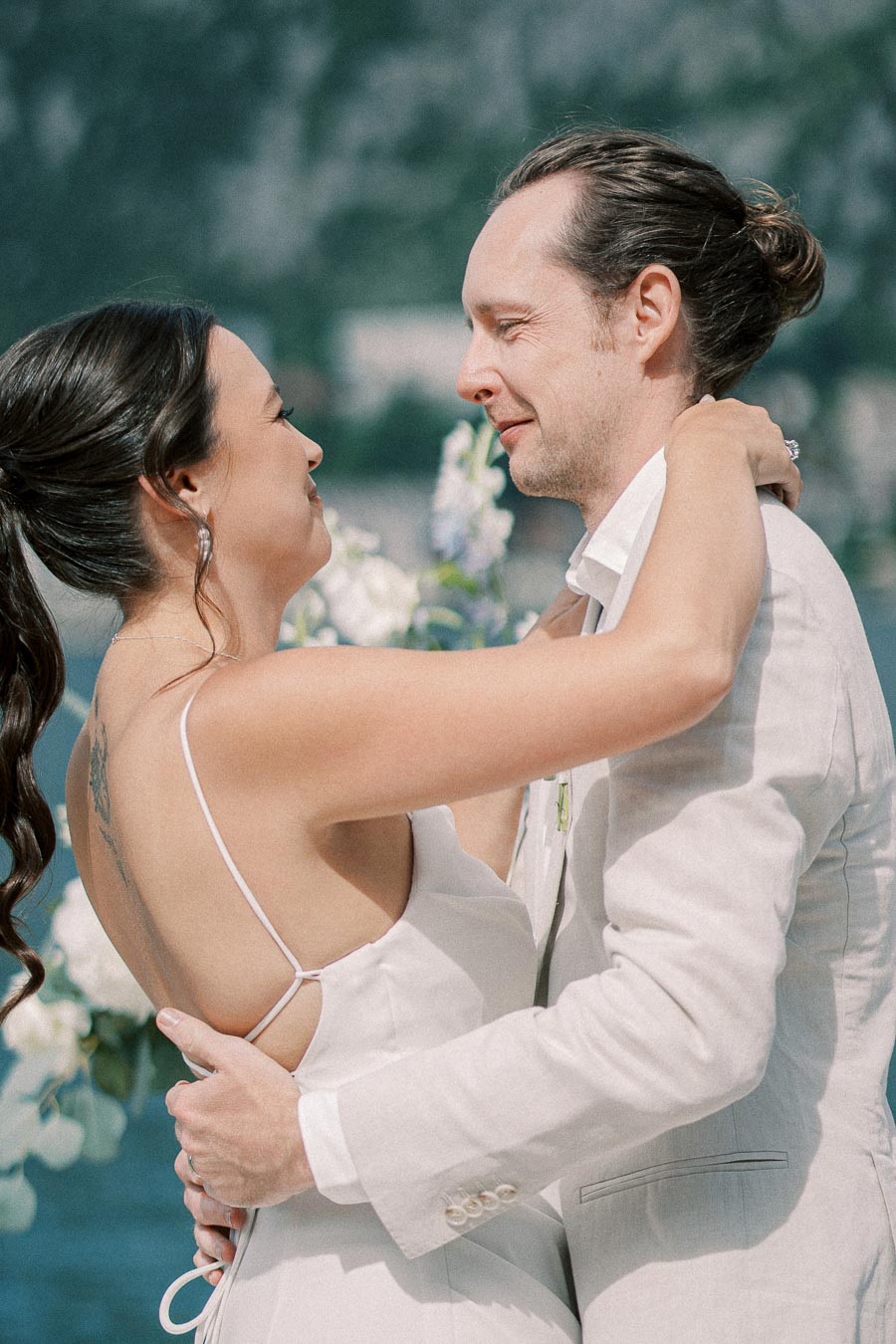 Couple embracing at an outdoor wedding, bride in white dress with floral decor in the background.