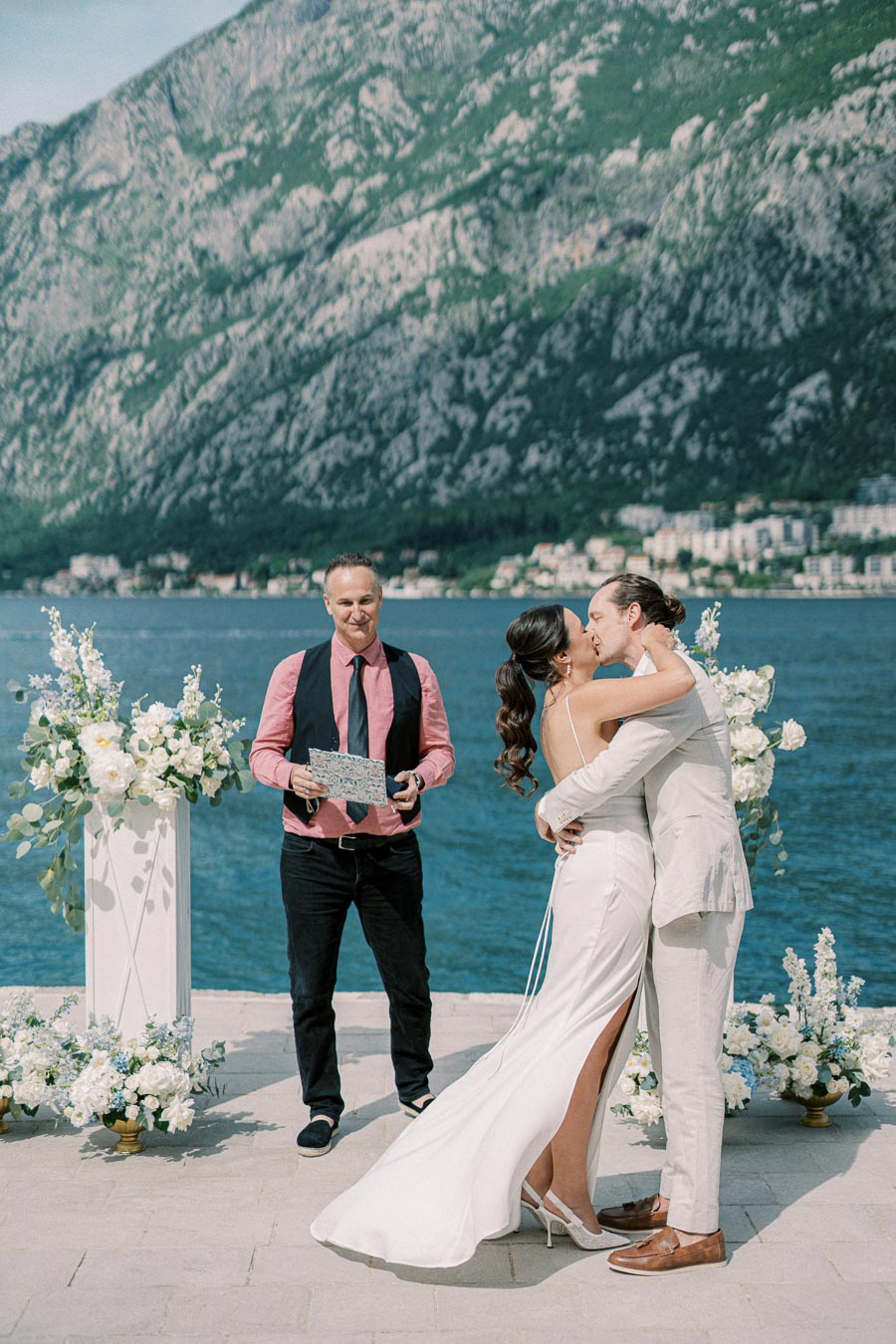 A couple kisses during an outdoor wedding ceremony by the sea, with a scenic backdrop of mountains and a celebrant smiling nearby. Floral arrangements decorate the venue under a clear, sunny sky.