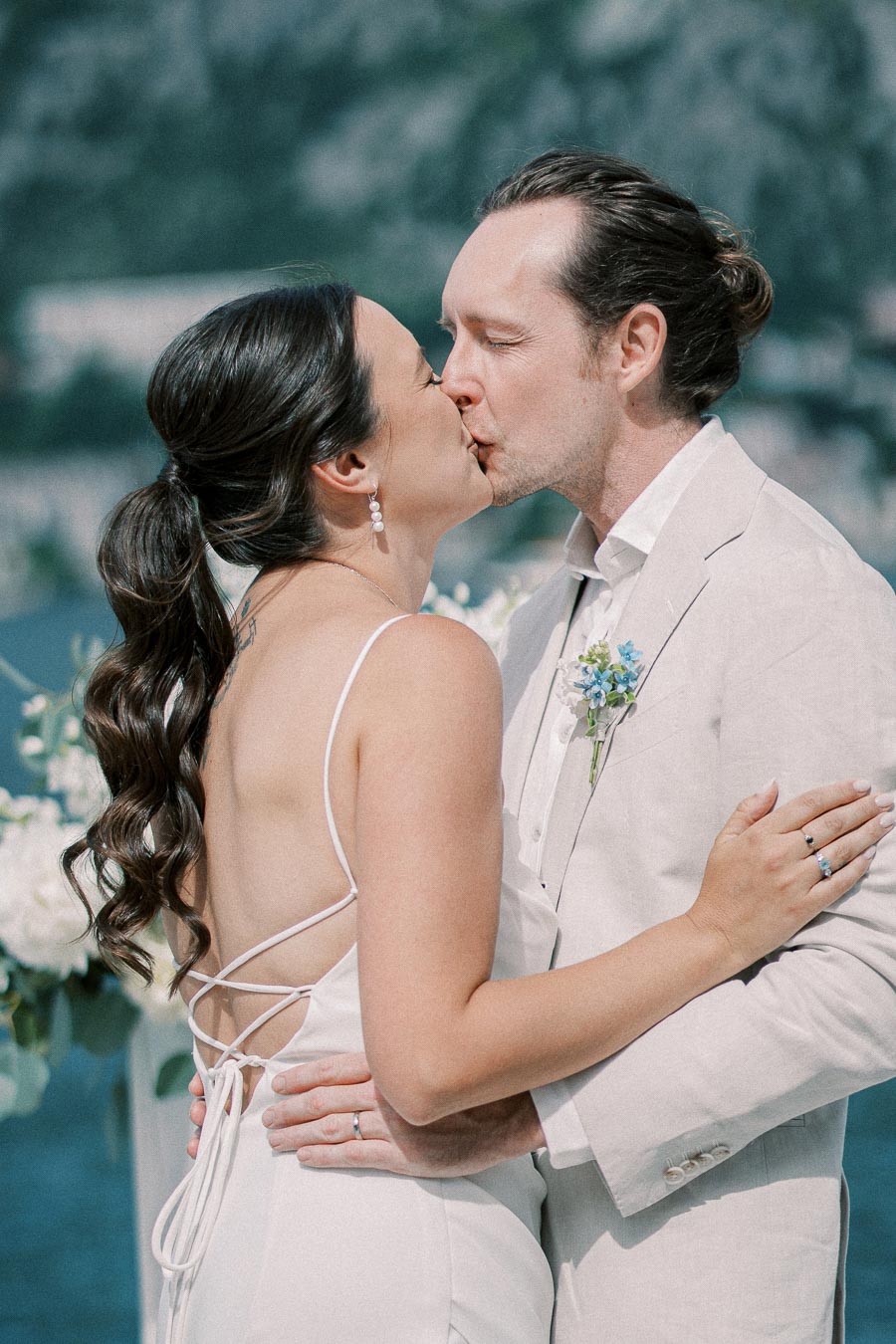 Romantic wedding couple kissing outdoors with scenic mountain and lake backdrop, groom in light suit and bride in elegant backless dress with floral bouquet.
