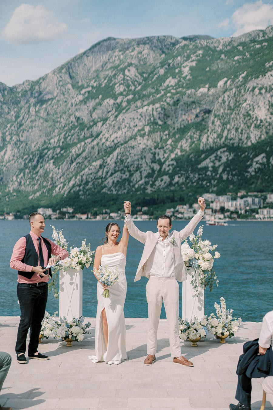 A newlywed couple celebrating after a waterfront wedding ceremony, with picturesque mountains and clear blue water in the background.