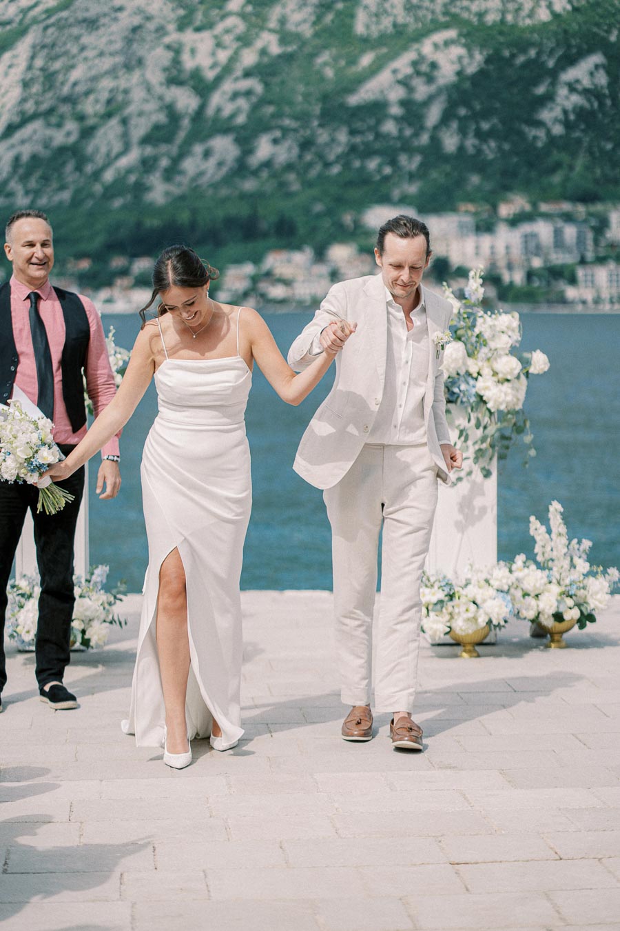Bride and groom holding hands and smiling outdoors at a lakeside wedding ceremony, surrounded by floral decorations and scenic mountains in the background.