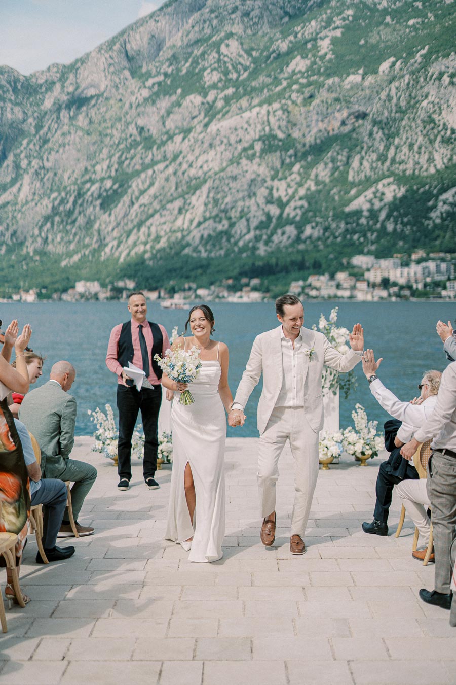A joyful couple holding hands and celebrating at an outdoor wedding ceremony by a picturesque lake, set against a backdrop of majestic mountains and applauding guests.