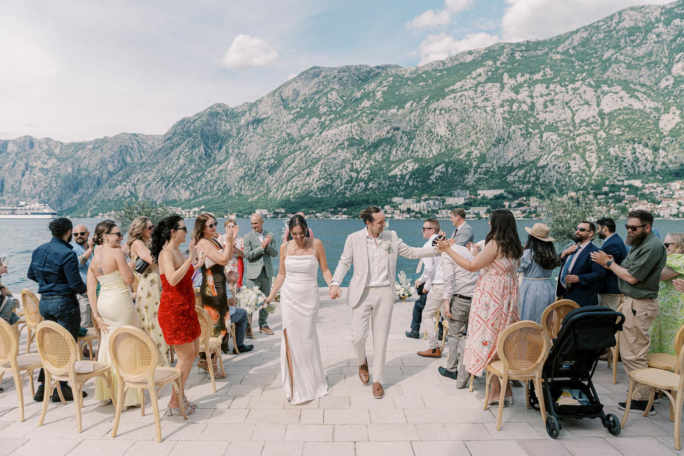 Beachfront wedding ceremony with a happy couple holding hands, surrounded by applauding guests, set against a backdrop of stunning mountains and a clear blue sky.