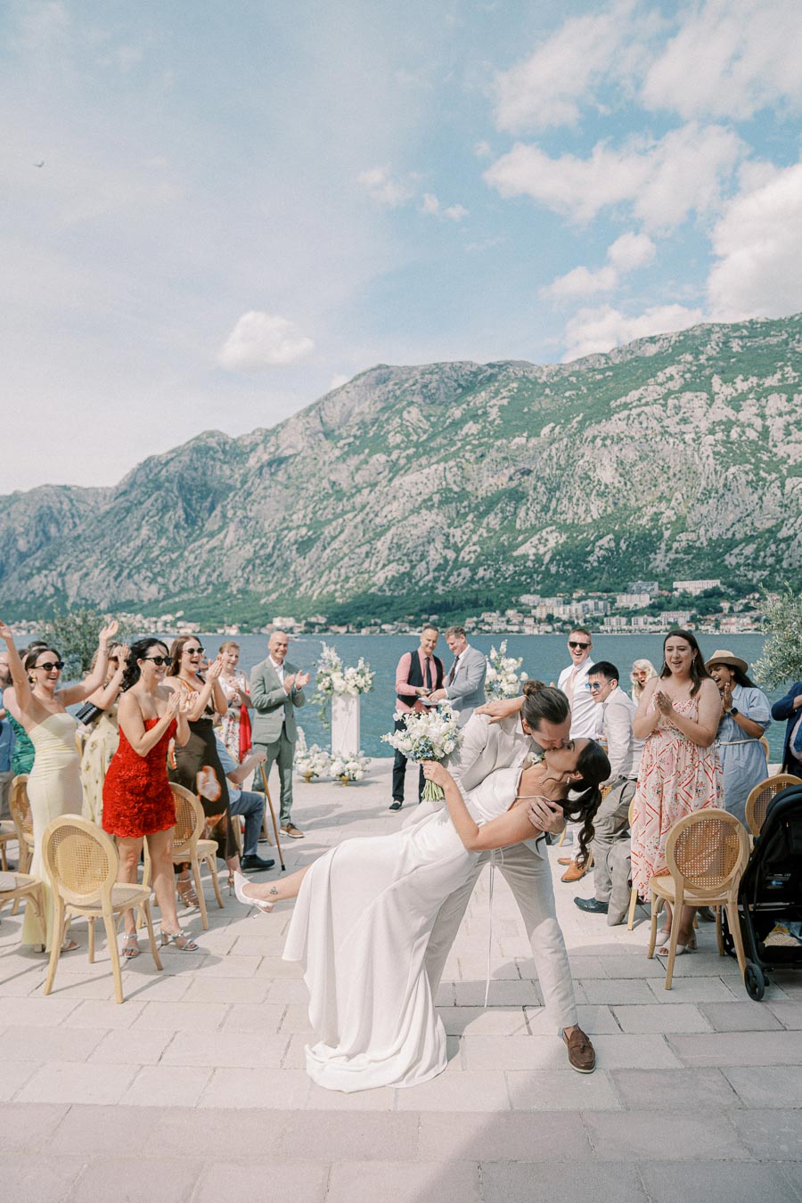 A bride and groom share a romantic kiss during their outdoor wedding ceremony by a picturesque lake, surrounded by joyous guests clapping and cheering, set against a backdrop of stunning mountains and a clear blue sky.