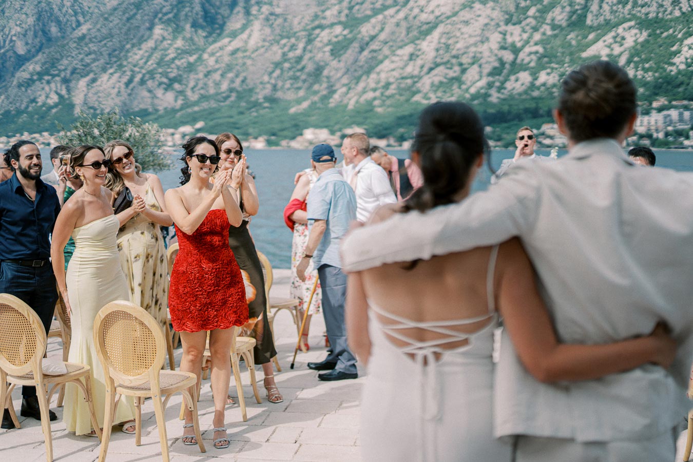 A joyful wedding ceremony with a couple embracing as guests applaud against a scenic backdrop of mountains and a lake.