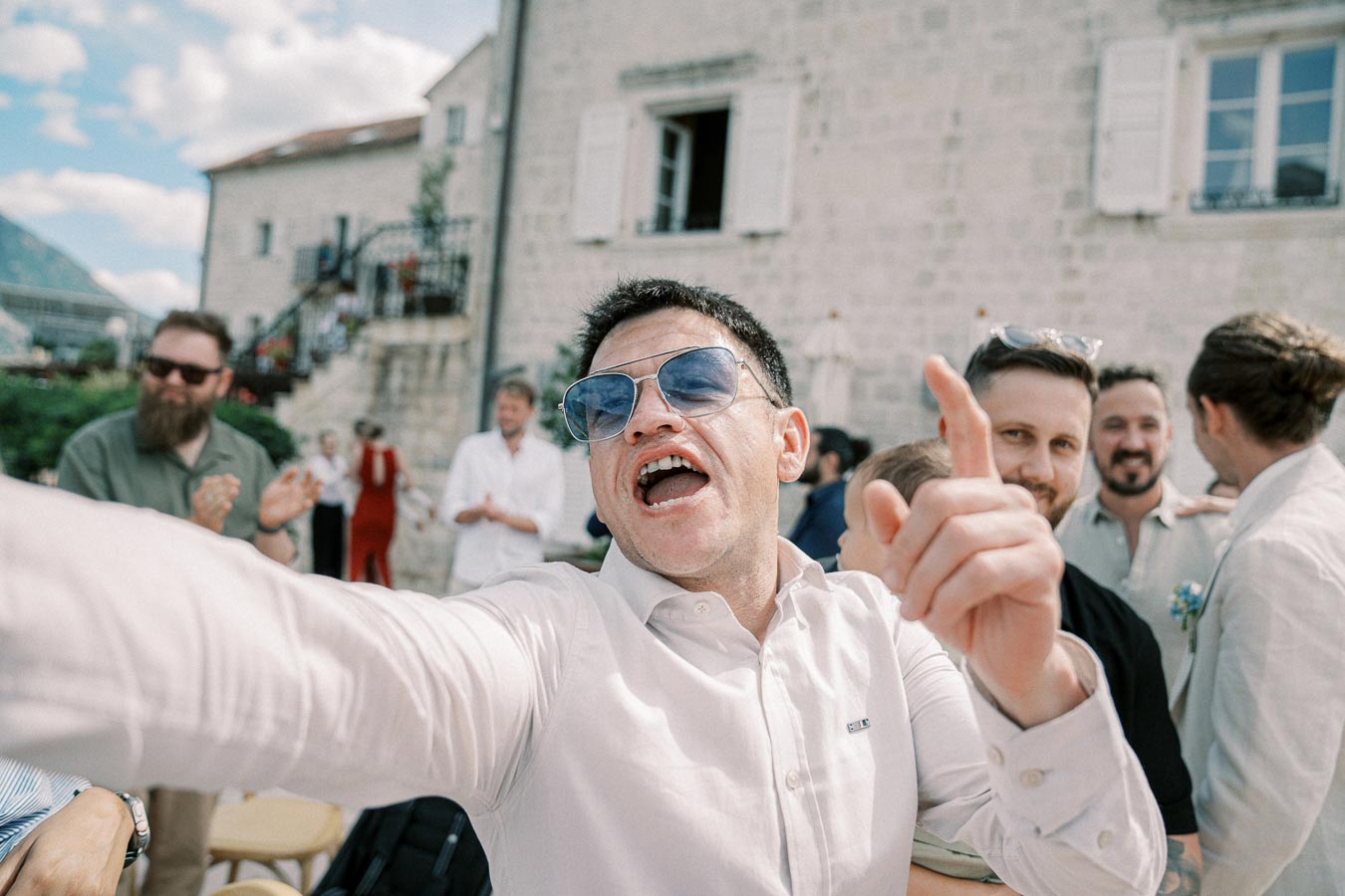 A man wearing blue sunglasses and a white shirt enthusiastically points and sings at an outdoor gathering, with a group of people smiling and enjoying the event in the background. Stone building and blue sky add to the lively atmosphere.