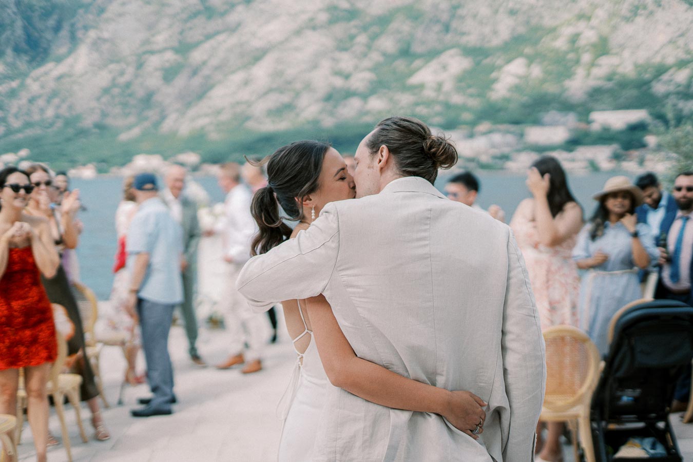 Couple kissing during outdoor wedding reception by a scenic mountain lake, surrounded by applauding guests and a picturesque background.