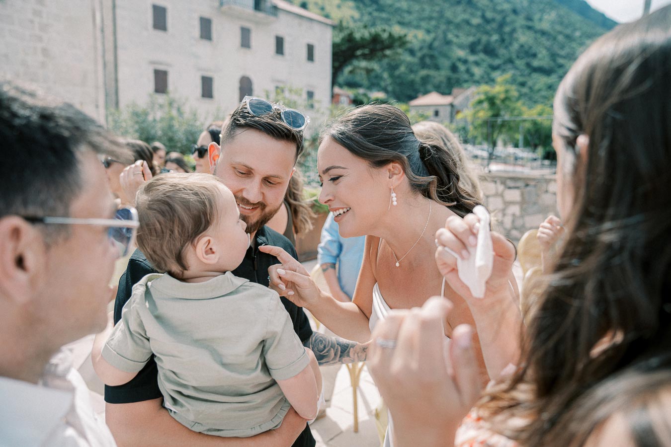 People enjoying an outdoor celebration, with a woman smiling at a baby held by a man, surrounded by a cheerful crowd and scenic landscape background.