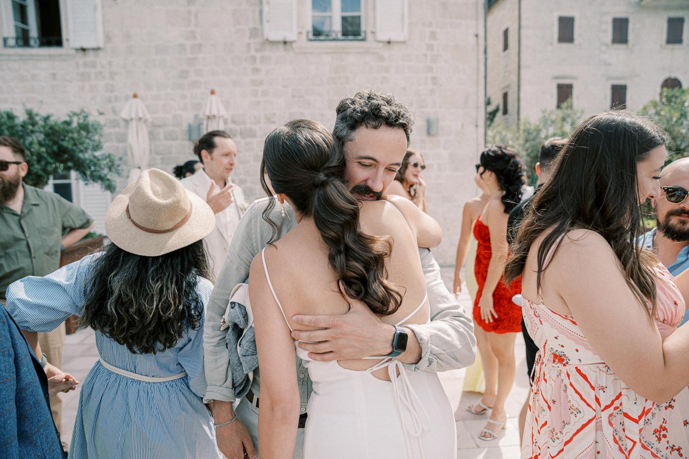 A group of people at an outdoor event, with a man and woman hugging warmly in the foreground. The woman is in a white dress, and several attendees are socializing in summer attire. The setting appears to be a sunny courtyard with stone buildings in the background.