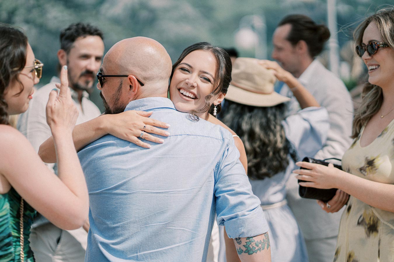 Group of friends enjoying a sunny outdoor gathering, with a smiling woman embracing a man in a light blue shirt, surrounded by others socializing and taking photos.