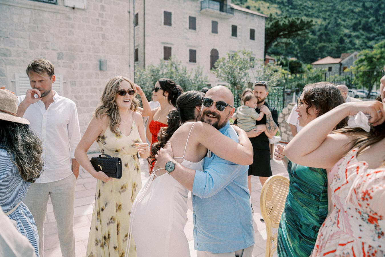 A group of people celebrating outdoors at a summer event, with a man in sunglasses hugging a woman in a white dress, surrounded by others in colorful attire against a backdrop of charming stone buildings and greenery.