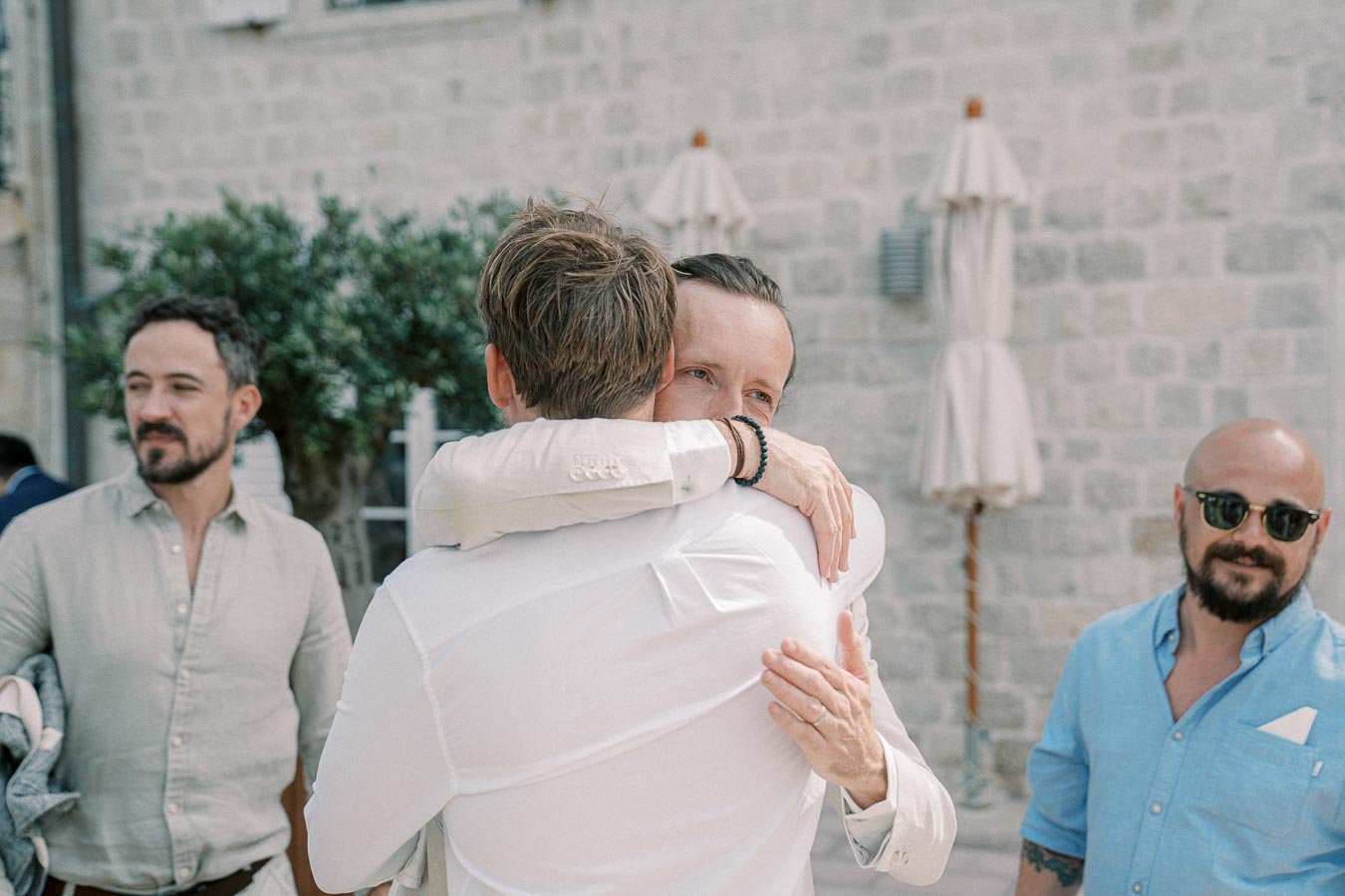 Group of men embracing and chatting in an outdoor setting with stone wall background, featuring two men hugging warmly and another smiling nearby, conveying friendship and celebration.