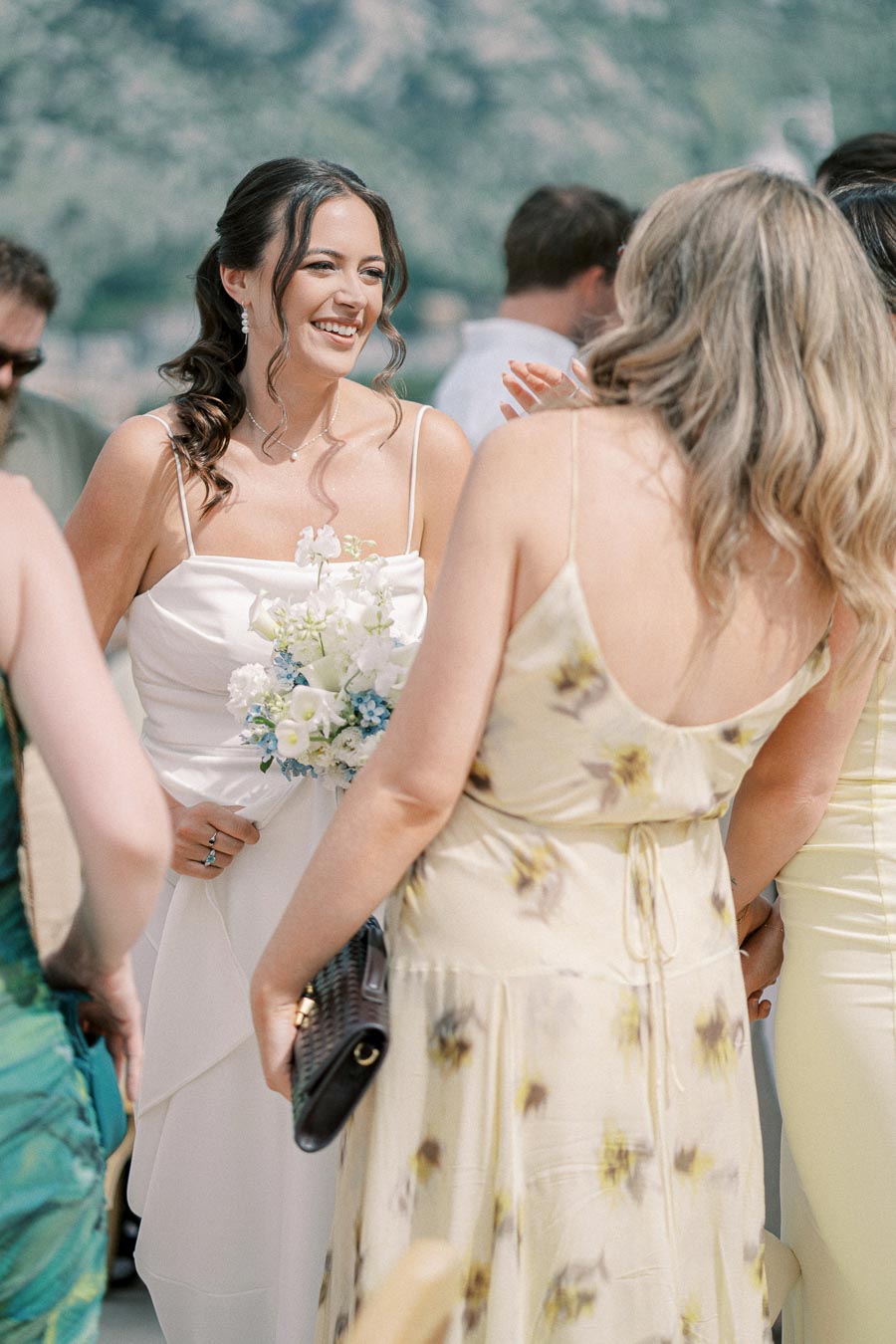 A smiling bride holding a bouquet of white and blue flowers, surrounded by guests in summer dresses at an outdoor wedding with a mountainous background.