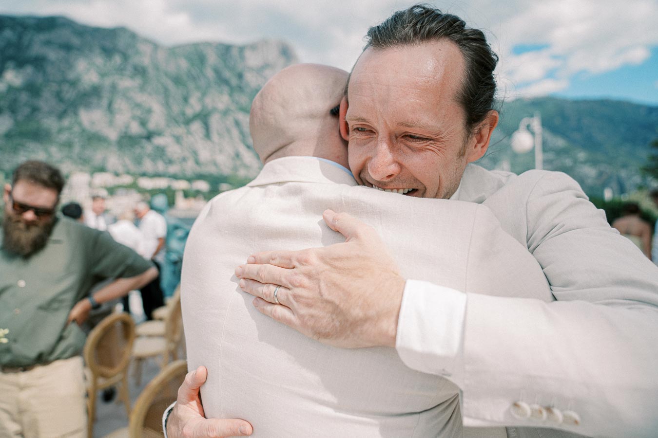 Two men in light-colored suits embracing warmly outdoors with a scenic mountain backdrop, capturing a joyful and celebratory moment.