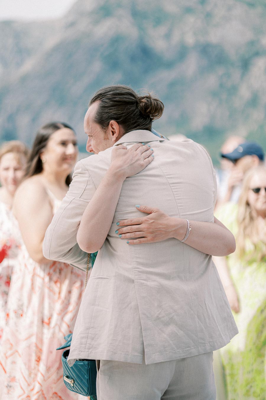 Two people embracing at an outdoor gathering, surrounded by smiling individuals in colorful summer attire, with mountains in the background on a sunny day.