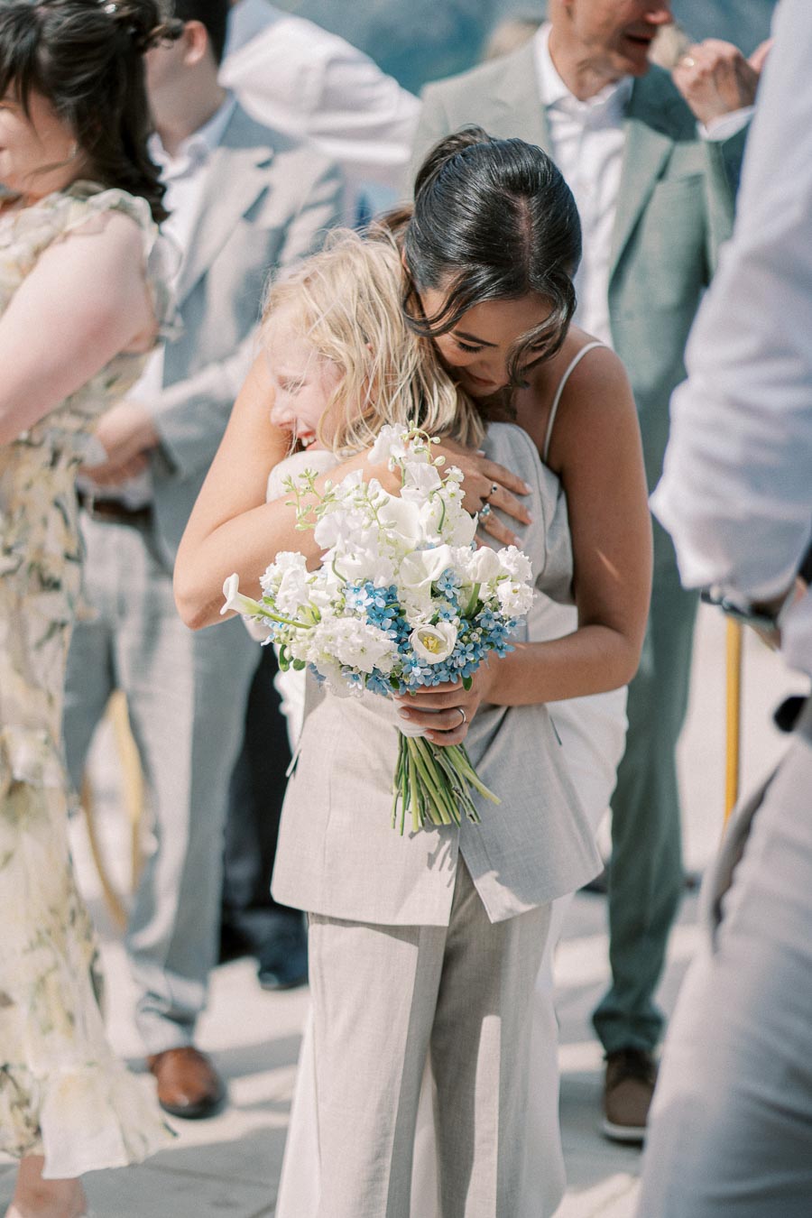 A bride embracing a child at a wedding ceremony, holding a bouquet of white and blue flowers, surrounded by guests in elegant attire.