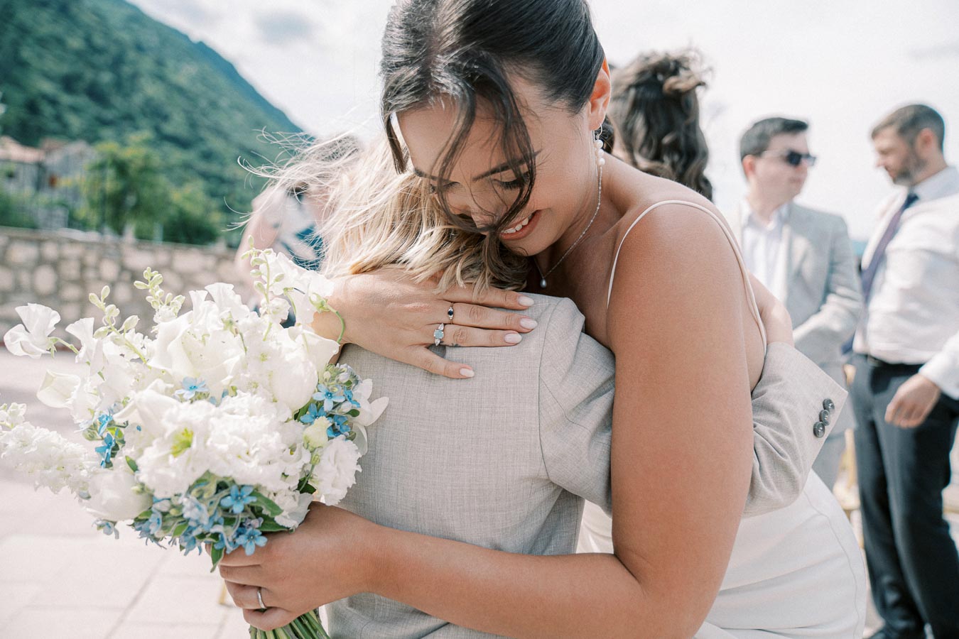 Bride embracing guest with a bouquet of white and blue flowers against a scenic outdoor background at a wedding ceremony.