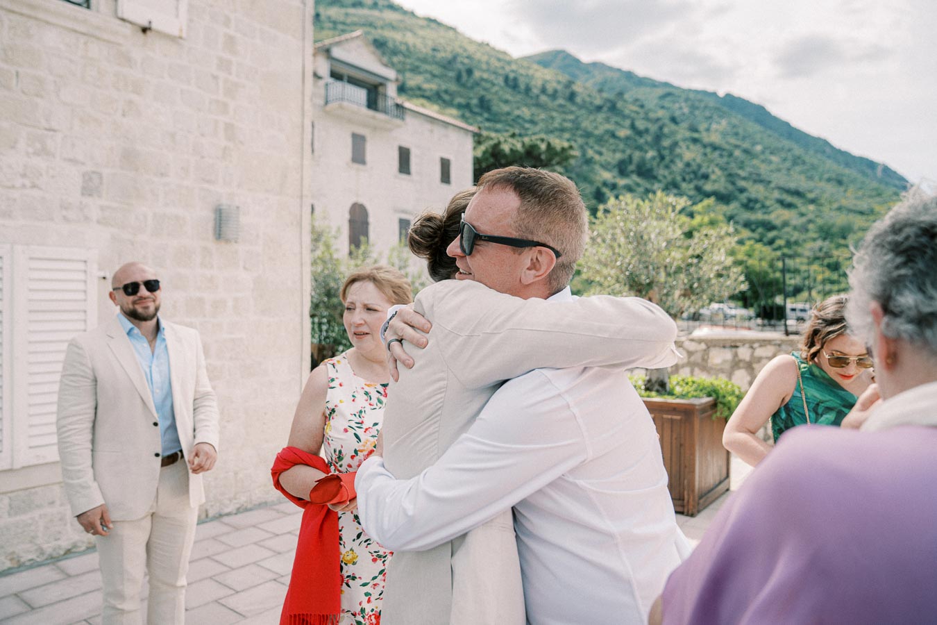A group of people gathered outdoors on a sunny day, with two individuals embracing in a hug. Others stand around them, wearing summer attire. The setting includes stone buildings and greenery in the background, suggesting a warm, scenic location.