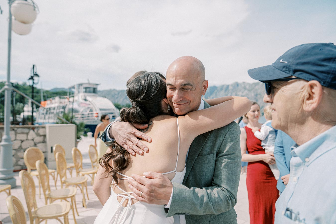 A joyful outdoor wedding celebration by a picturesque lake, where a bride embraces a smiling guest, surrounded by attendees and scenic mountains in the background.