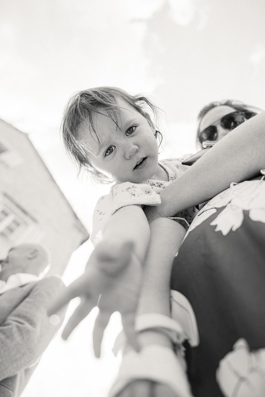 Black and white image of a small child reaching towards the camera, supported by an adult in sunglasses, with a blurred building in the background.